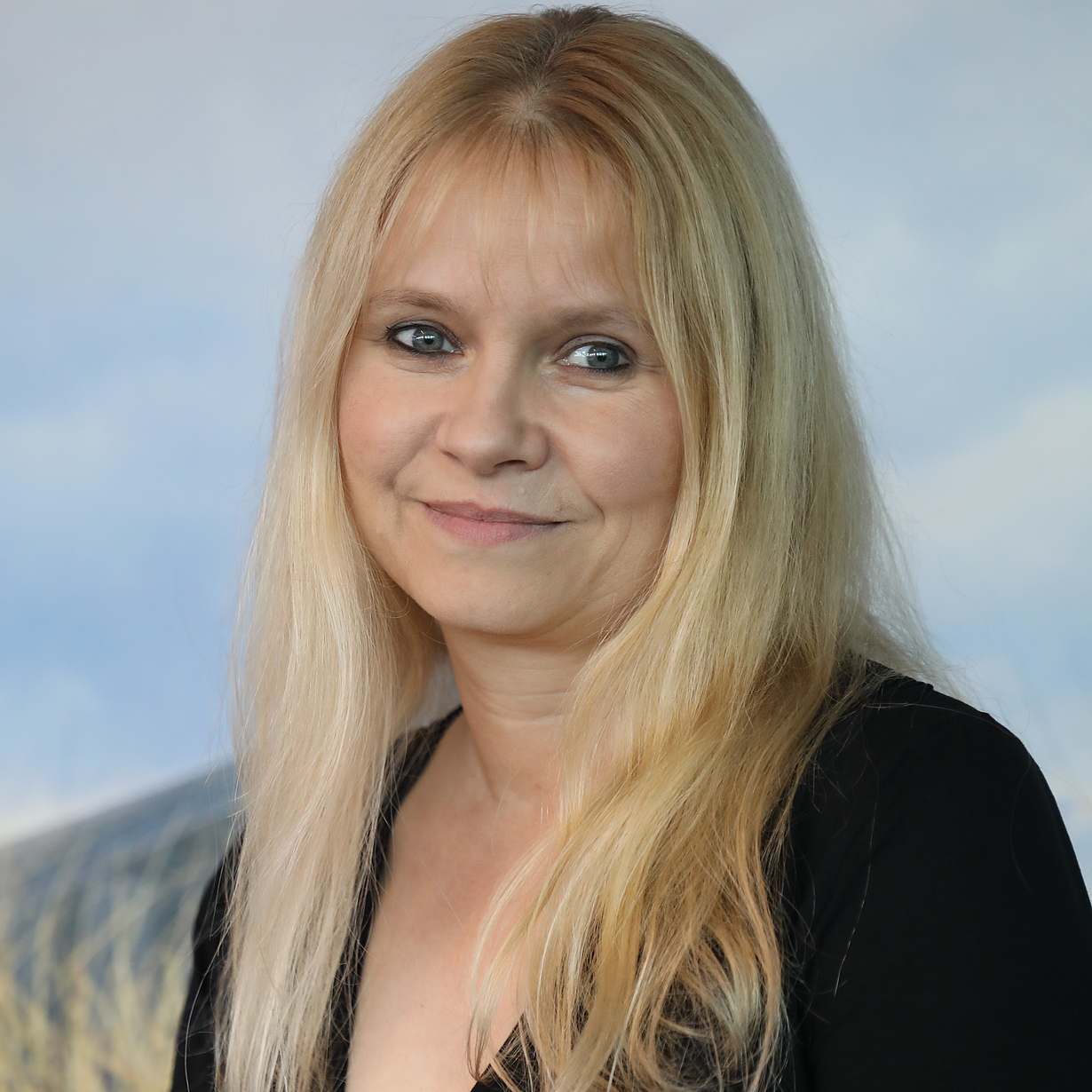 Eine Frau mit langen, blonden Haaren und einem freundlichen Lächeln steht vor einem sanften, natürlichen Hintergrund. Sie trägt ein schwarzes Oberteil und sieht in die Kamera. Ihr Ausdruck wirkt freundlich und offen.