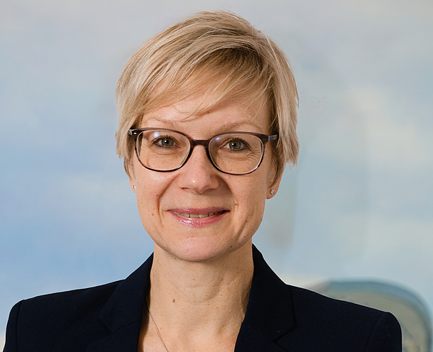 Ein Porträt einer lächelnden Frau mit blonden, kurzen Haaren und Brille, die ein gestreiftes Oberteil unter einem dunklen Blazer trägt. Der Hintergrund zeigt unscharfe, hellblaue und gelbliche Farben.